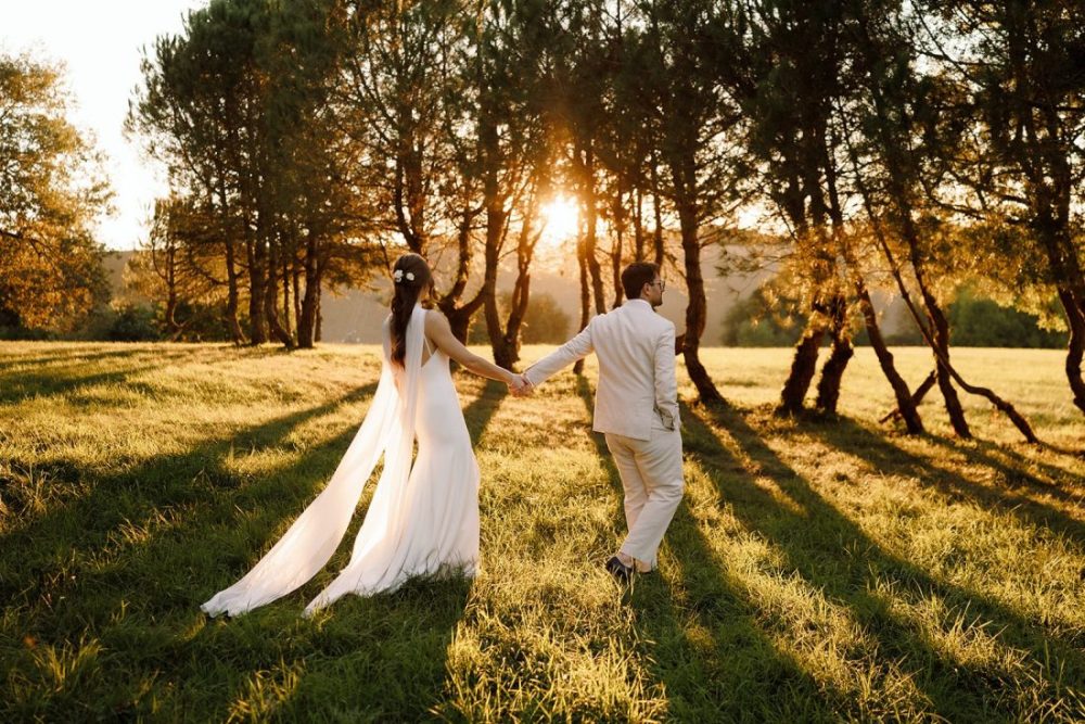 The groom leads his bride through the trees with the sunlight casting long shadows